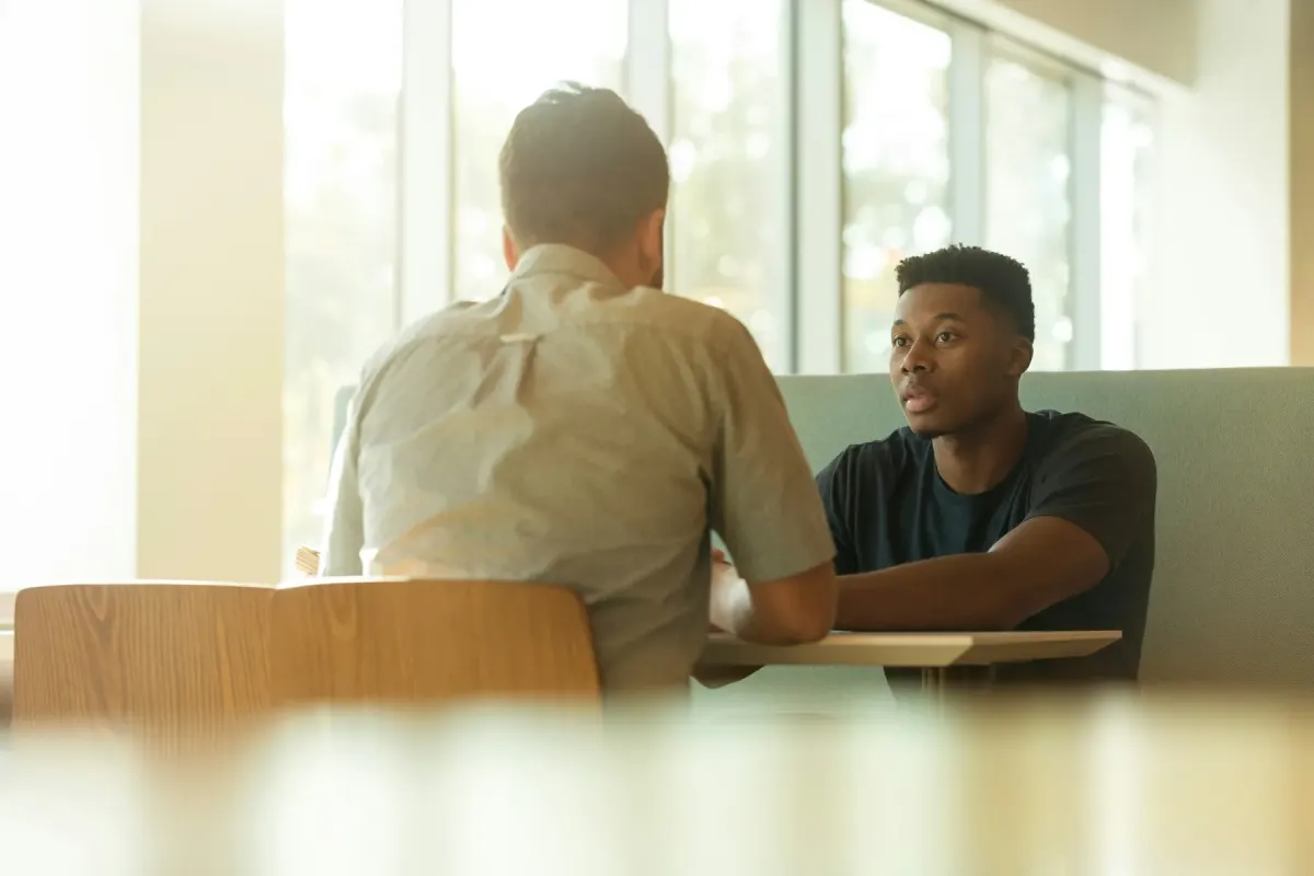 Two people in conversation across a small table in a sunlit room